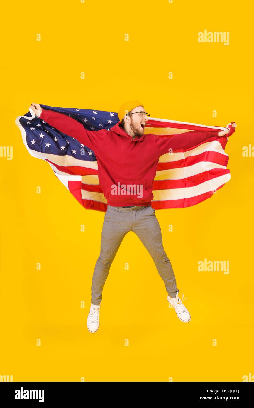 Heureux 4th juillet, un beau jeune homme célèbre la nuit jumpante avec le drapeau des États-Unis derrière son dos isolé sur fond jaune. La liberté est dans votre vie. Jeune homme joyeux avec drapeau américain. Banque D'Images