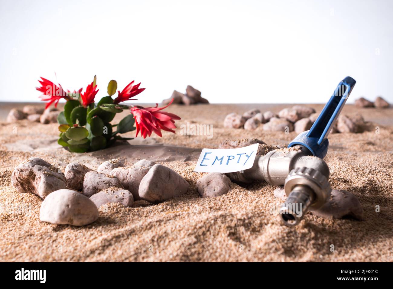 Gros plan d'un robinet ouvert et sec enterré par du sable désertique avec une plante succulente sur fond sec et blanc de ciel Banque D'Images