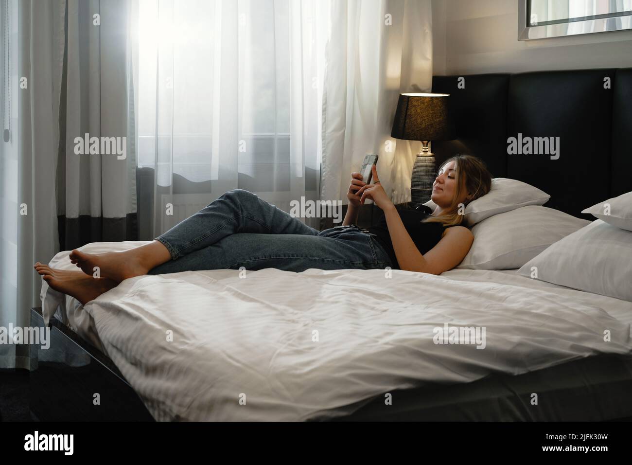 Jeune femme détendue dans le lit de la chambre d'hôtel regardant le smartphone, la planification de la journée, le chat, la lecture. Femme qui se repose dans un hôtel moderne pendant son voyage Banque D'Images