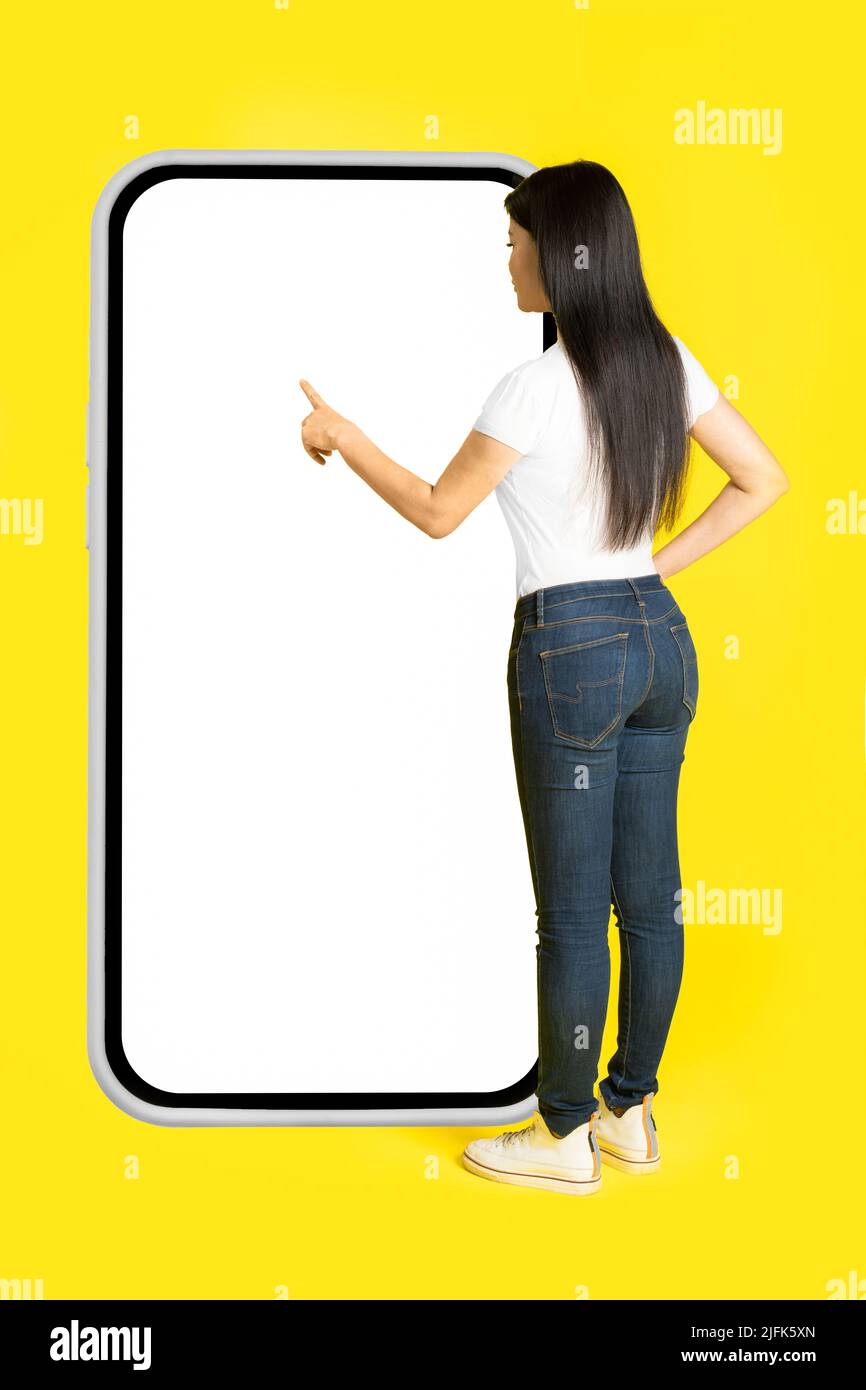 Charmante femme asiatique d'âge moyen avec un téléphone dans les mains montrant un écran blanc adossé sur un immense smartphone avec un écran blanc portant un style décontracté isolé sur fond jaune. Maquette d'espace libre. Banque D'Images