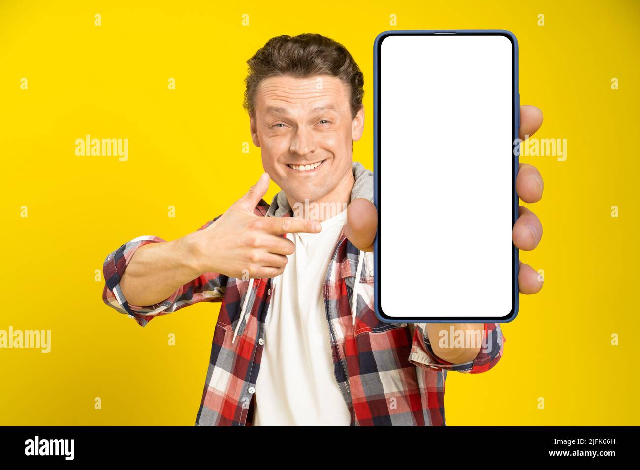 Charmant homme heureux pointant vers un smartphone avec écran blanc vide, portant une chemise à carreaux rouge et un Jean écran de téléphone portable maquette isolée sur fond jaune. Publicité pour applications mobiles. Banque D'Images