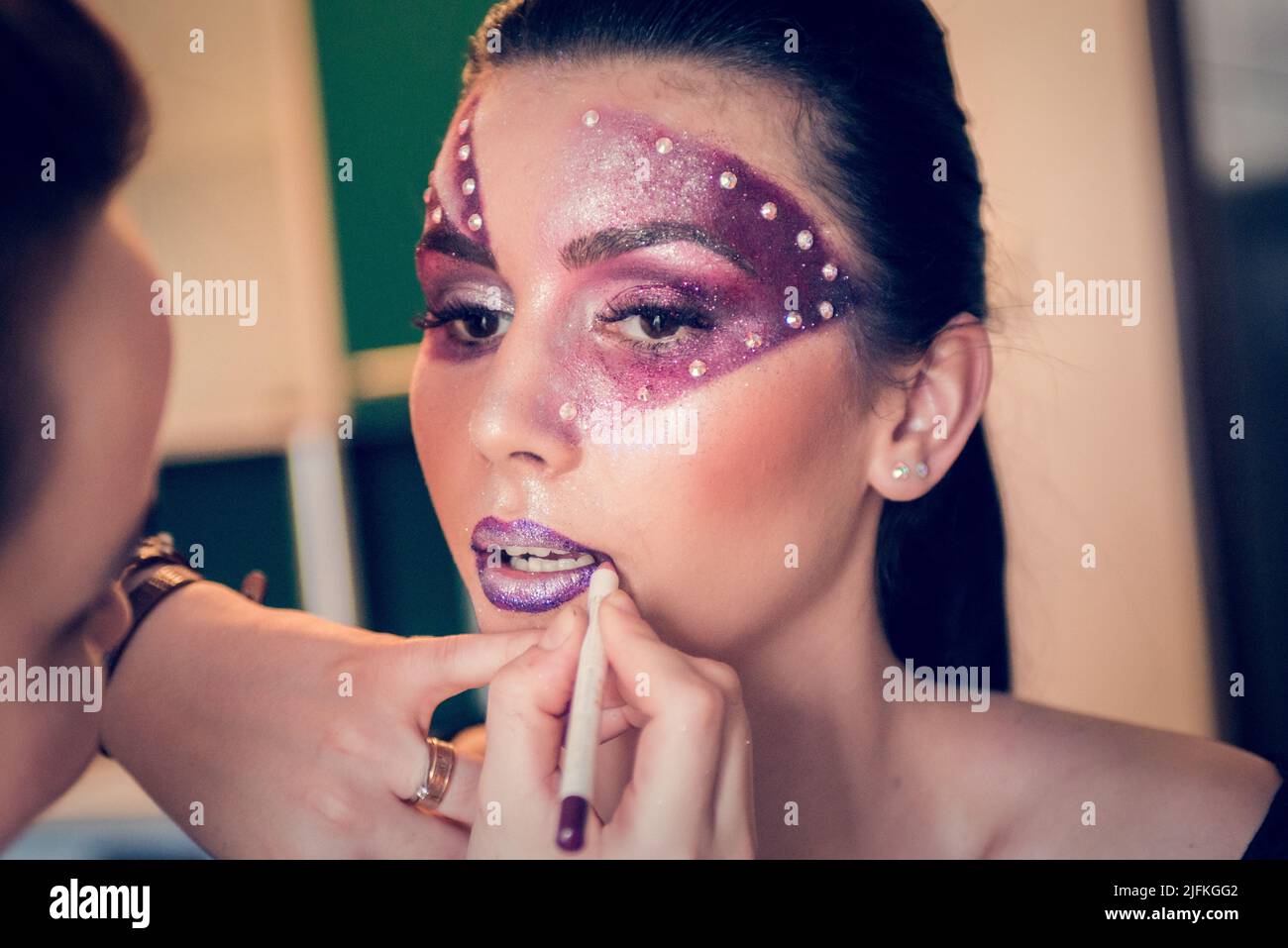 Après avoir peint le visage du modèle avec du violet et des cristaux, l'artiste de maquillage façonne ses lèvres avec un stylo de contour violet Banque D'Images