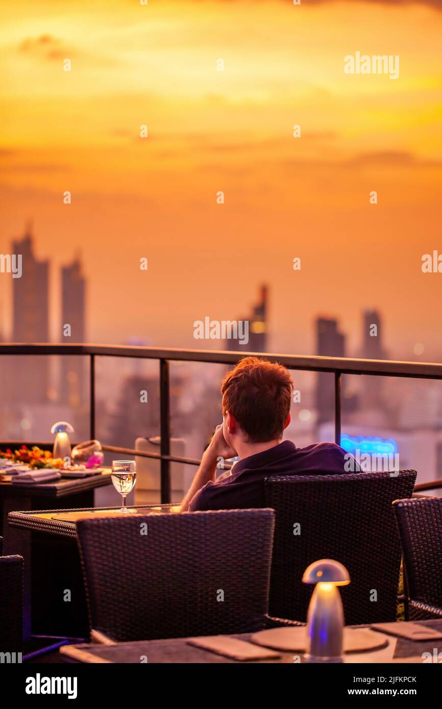 Un jeune homme avec un verre de vin se reposer dans un luxueux restaurant sur le toit, en regardant le coucher du soleil sur le ciel orange. Homme avec un cocktail sur la terrasse du bar SKY Banque D'Images