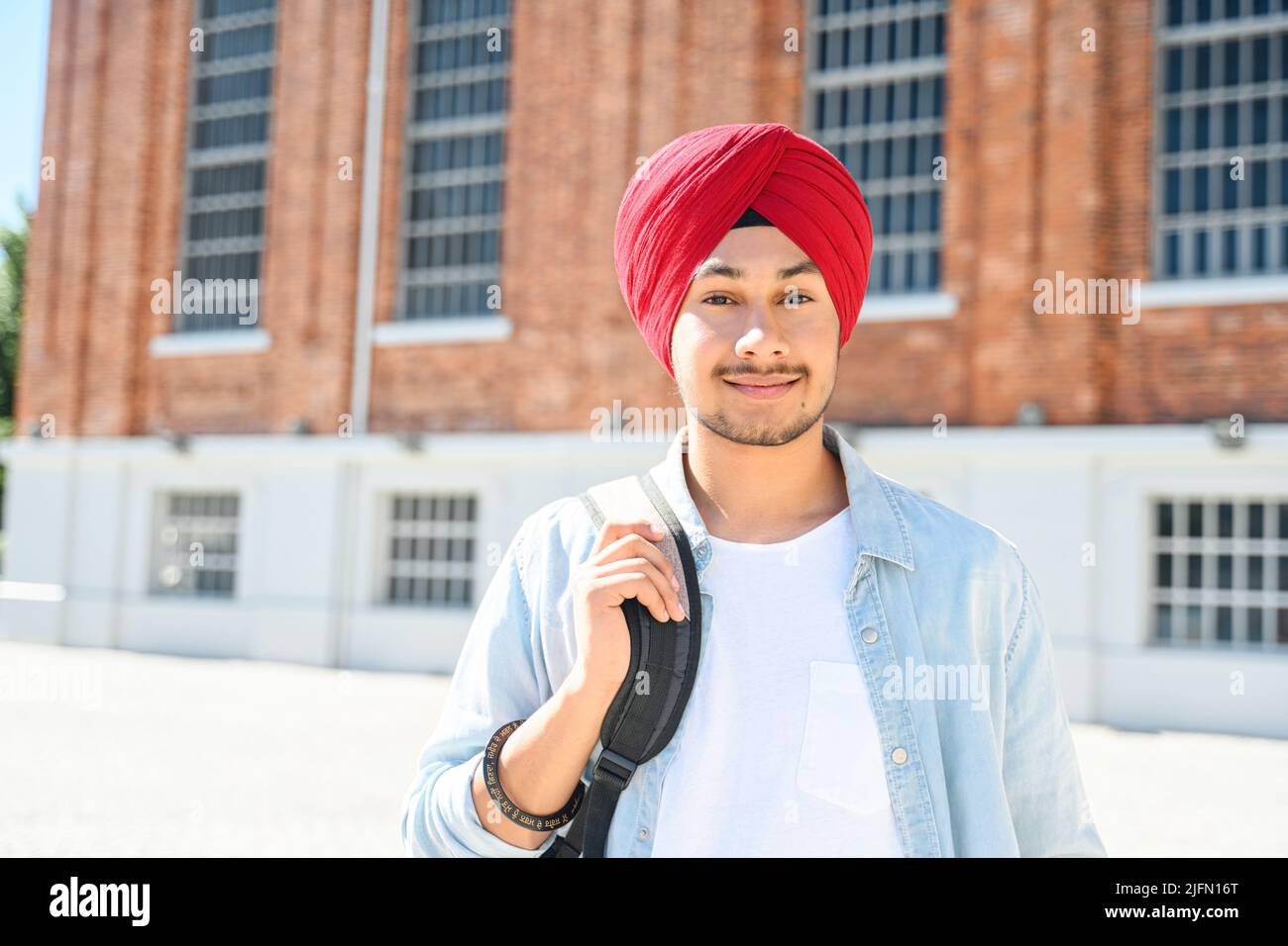 Portrait d'un jeune étudiant indien heureux portant un pagg traditionnel rouge turban et une chemise décontractée sur le campus, un homme hindou souriant optimiste avec sac à dos debout à l'extérieur près de l'université Banque D'Images
