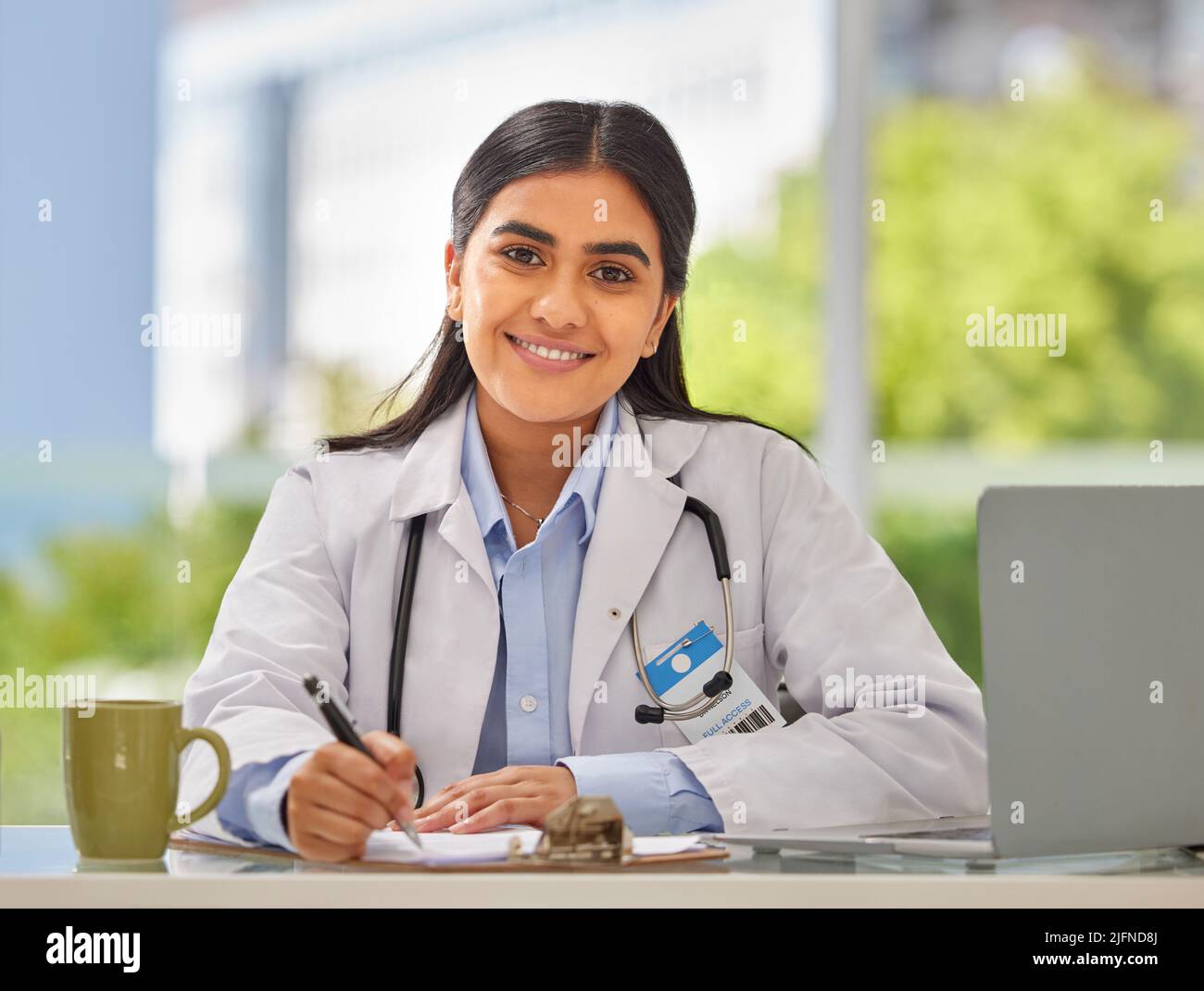 Portrait d'un jeune médecin indien portant un stéthoscope assis dans un bureau écrivant une ordonnance tout en étant assis à son bureau. Femme souriante gp or Banque D'Images