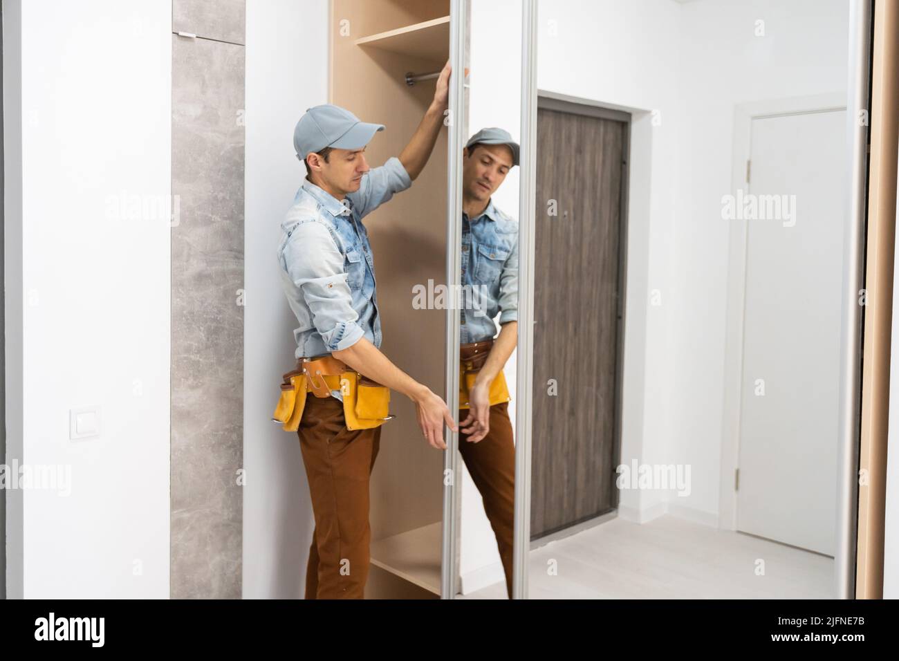 L'assembleur de mobilier employé en t-shirt blanc et combinaison-pantalon vérifie la porte coulissante en bois de l'armoire moderne avec miroir. Banque D'Images