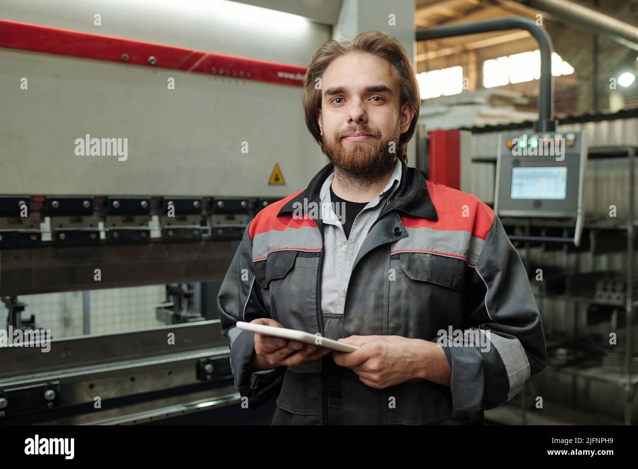 Jeune ingénieur confiant avec une tablette debout devant la caméra dans l'atelier de l'usine industrielle et travaillant sur le net Banque D'Images