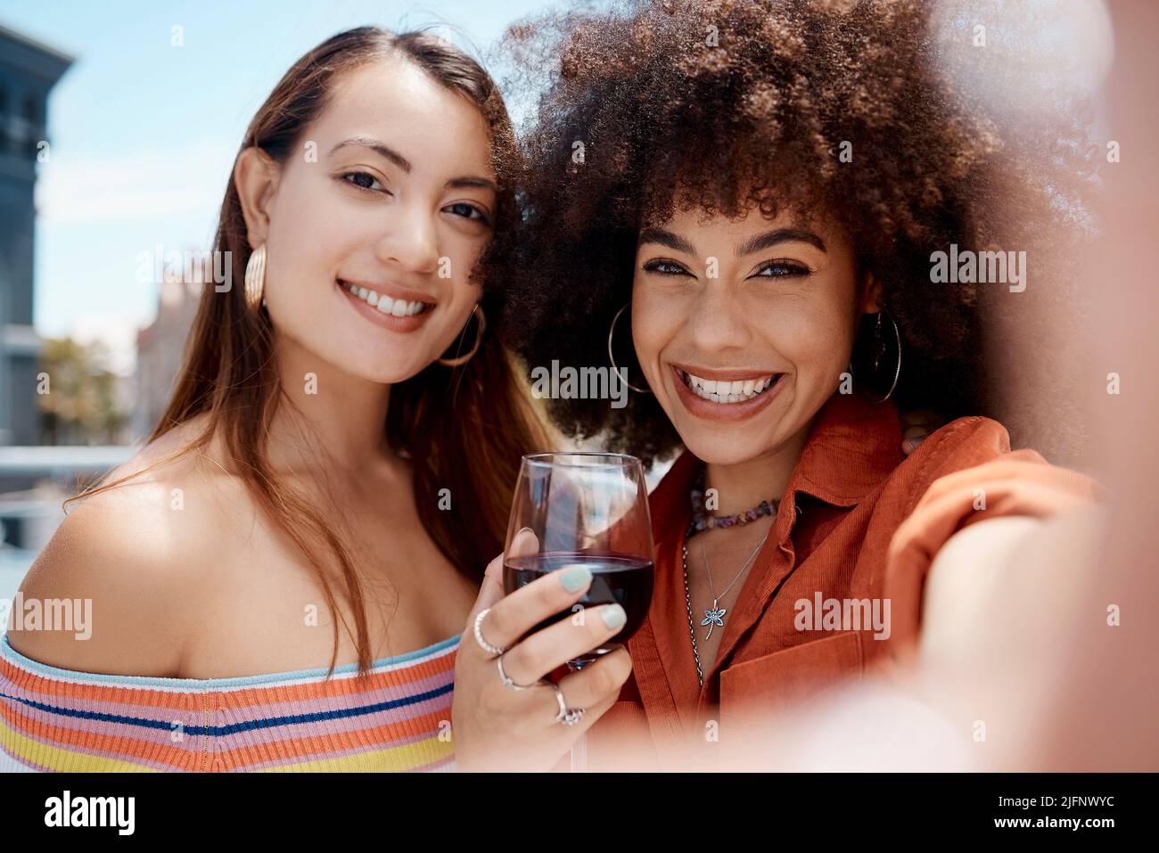 Deux belles femmes de course mixte souriant prenant un selfie tout en dégustant un verre de vin rouge pendant une fête à l'extérieur. Femme hispanique avec afro frais Banque D'Images