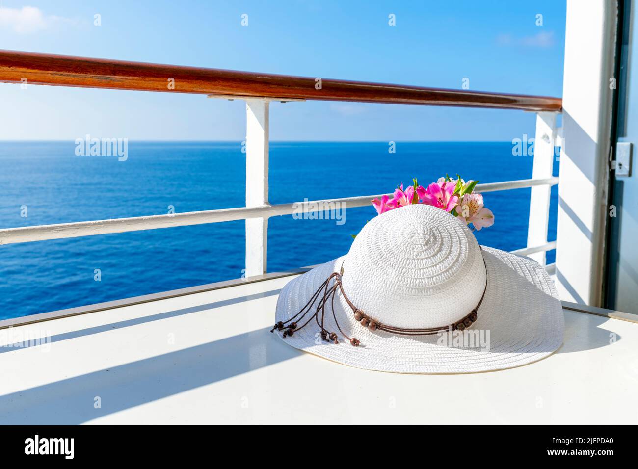 Un chapeau de soleil blanc pour femme se trouve à côté d'un petit bouquet de fleurs sur le balcon d'un bateau de croisière avec vue sur la mer bleue pendant une journée d'été. Banque D'Images