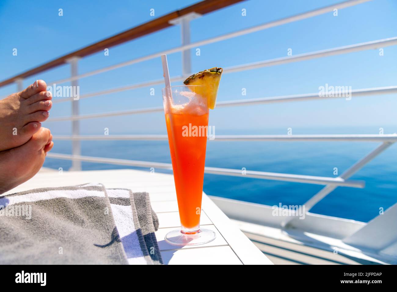 Une femme repose ses pieds sur une table sur le pont supérieur d'un bateau de croisière en mer avec une boisson colorée à l'ananas lors d'une chaude journée d'été sur l'océan. Banque D'Images