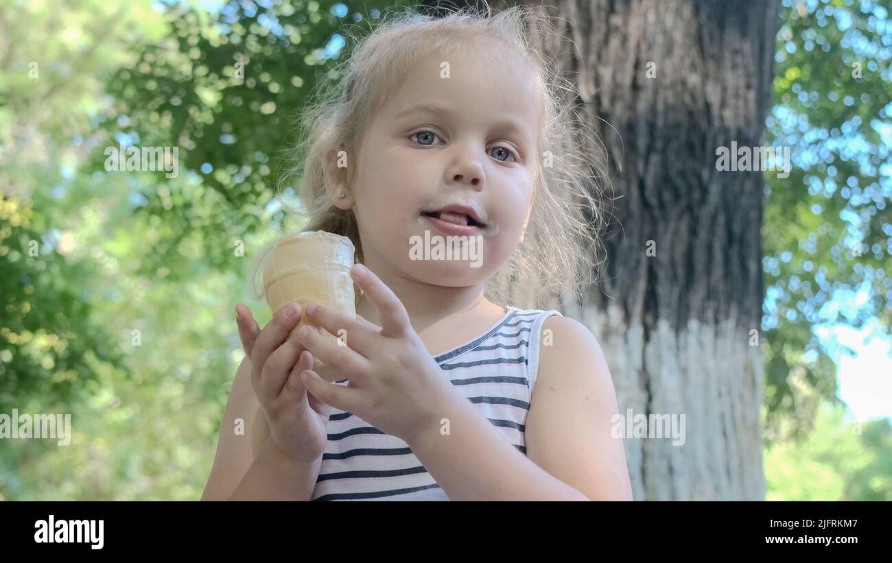 Une petite fille mignonne mange de la glace à l'extérieur. Portrait en gros plan d'une fille blonde assise sur le banc du parc et mangeant de la glace. Banque D'Images