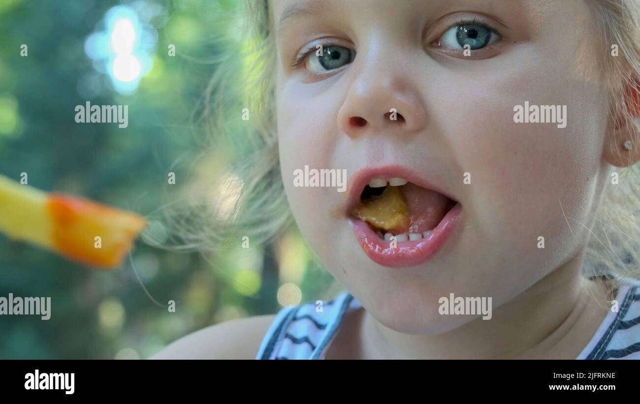 Une petite fille mange des frites. Gros plan de blonde fille prend des chips de pomme de terre avec ses mains et les essaie assis dans la rue café sur le parc. Banque D'Images