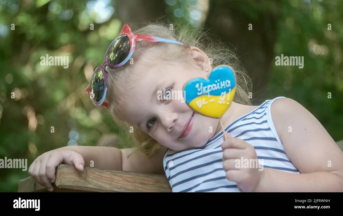 Odessa, Ukraine, Europe de l'est. 3rd juillet 2022. Petite fille blonde tient le pain d'épice dans les couleurs nationales ukrainiennes dans sa main, il dit ''l'Ukraine est moi'. Portrait en gros plan d'une fille assise sur le banc du parc avec des biscuits aux couleurs du drapeau ukrainien. Ralenti. Odessa Ukraine (Credit image: © Andrey Nekrasov/ZUMA Press Wire) Banque D'Images