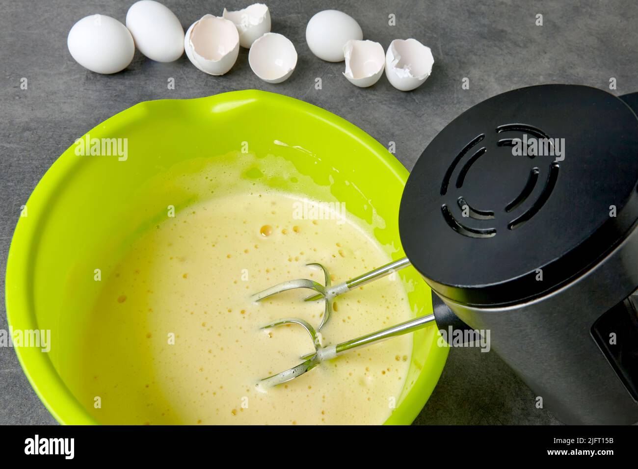 Battue les œufs avec du sucre dans un bol vert avec un batteur immergé dans un batteur et des coquilles d'œufs de poulet sur une table de cuisine en béton foncé. Création de la taille Banque D'Images