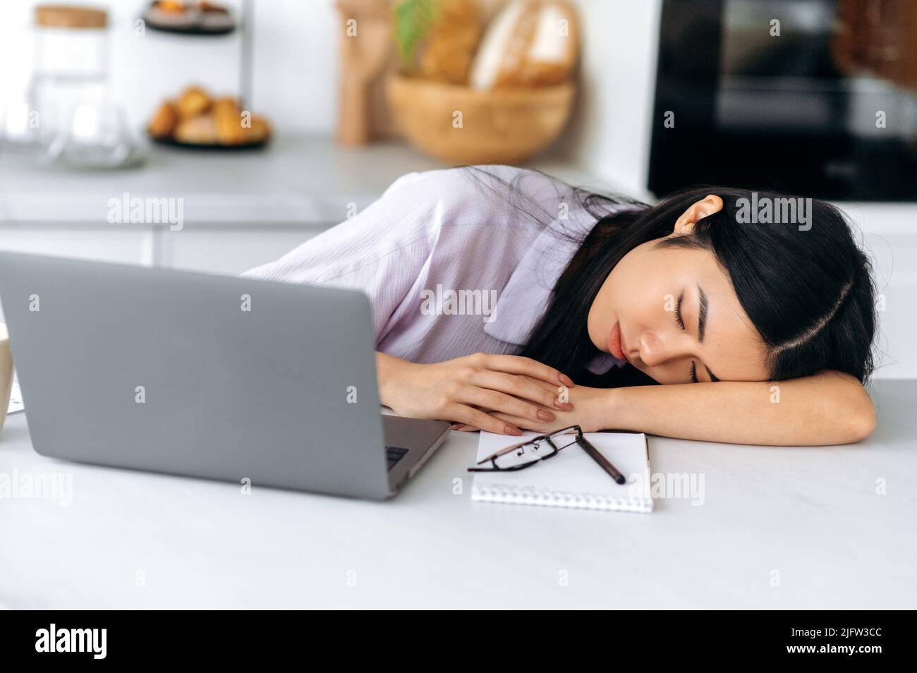 Une jeune femme d'origine chinoise somnolente s'endormit alors qu'elle était assise sur le lieu de travail près d'un ordinateur portable dans la cuisine, fatiguée d'un travail ennuyeux, surtraitée, souffrant de privation chronique de sommeil, a besoin de repos Banque D'Images
