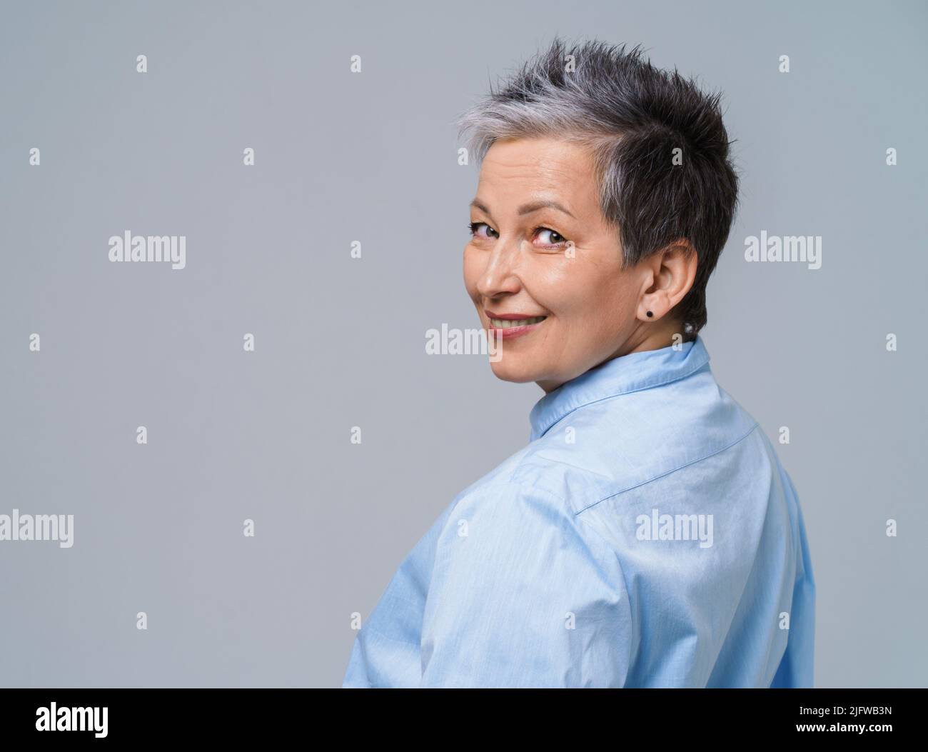 Cheveux gris mature femme posant debout dos demi-tour regardant par-dessus l'épaule à l'appareil photo et sourire portant une chemise bleue isolée sur fond blanc. Une beauté vieillie. Copier l'espace. Banque D'Images