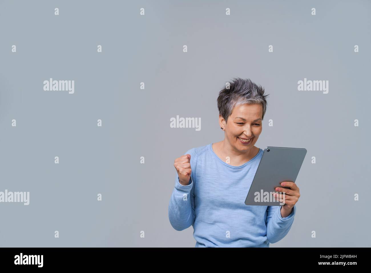Femme mûre et enthousiaste aux cheveux gris regardant les achats de Tablet pc, travaillant en ligne ou ayant un appel vidéo. Jolie femme en 50s portant un chemisier bleu isolé sur le gris. Des personnes et des technologies matures. Banque D'Images