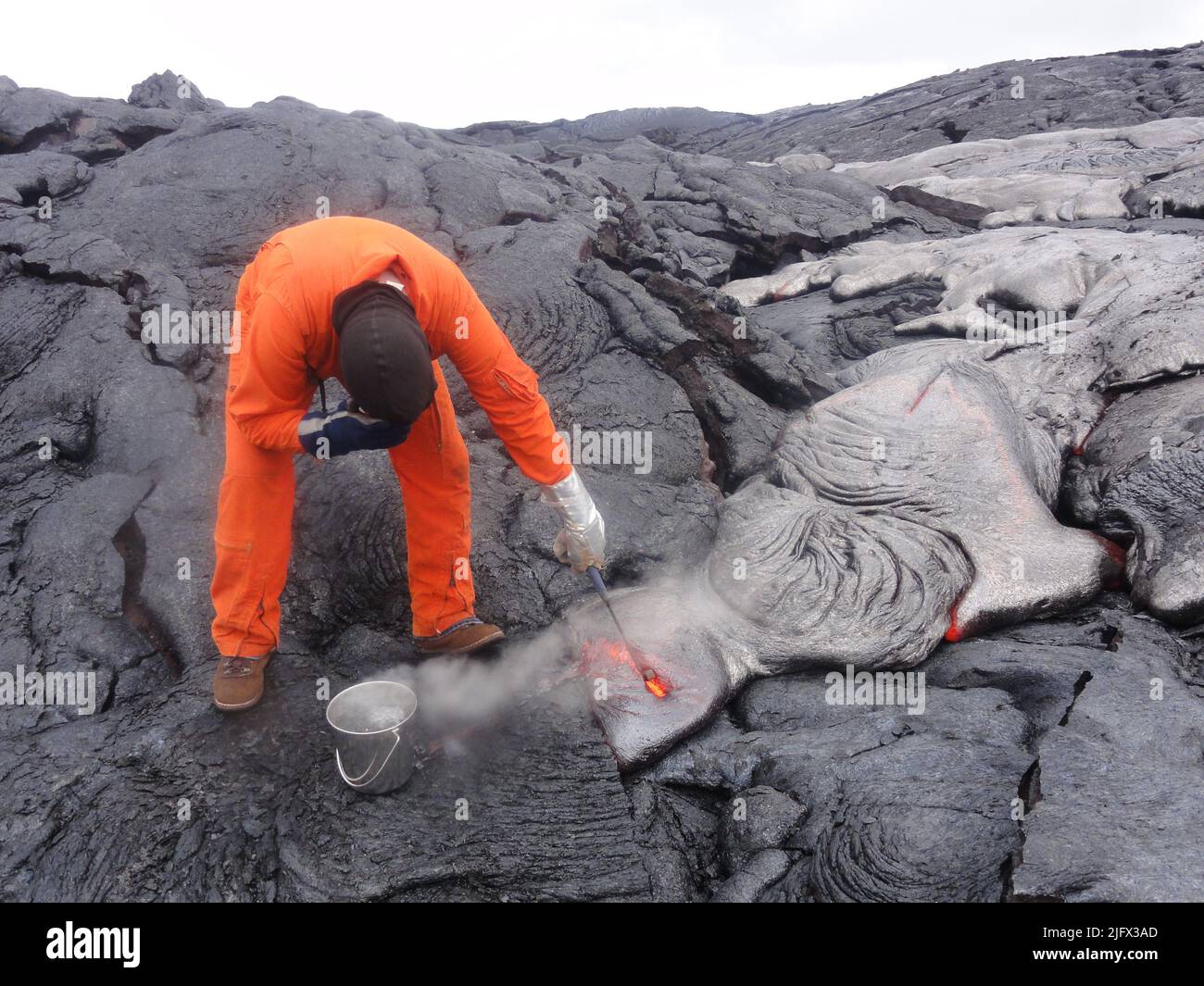 Un géologue de l'Observatoire du volcan hawaïen protège son visage de la chaleur intense alors qu'il plonge un marteau de roche dans un pied de p?hoehoe actif. Après avoir fait sortir la lave, elle est placée dans l'eau pour la refroidir. HVO collecte régulièrement des échantillons de lave pour l'analyse chimique, ce qui peut donner un aperçu des changements dans le système magmatique. Crédit : USGS Banque D'Images