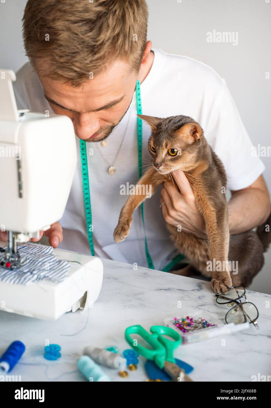 Couture de vêtements pour animaux de compagnie. Vêtements de tailleur pour chats. Un homme souriant est en train de coudre des vêtements pour son chat abyssinien. Le concept d'une petite entreprise de bricolage. Banque D'Images