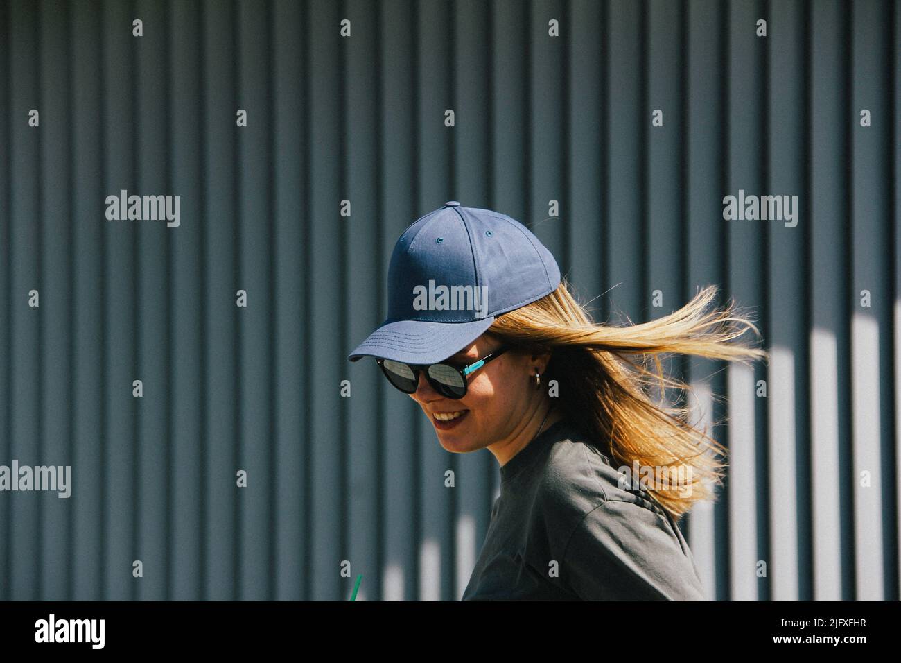 une fille dans une casquette et des lunettes de soleil sourit contre un mur gris nervuré Banque D'Images