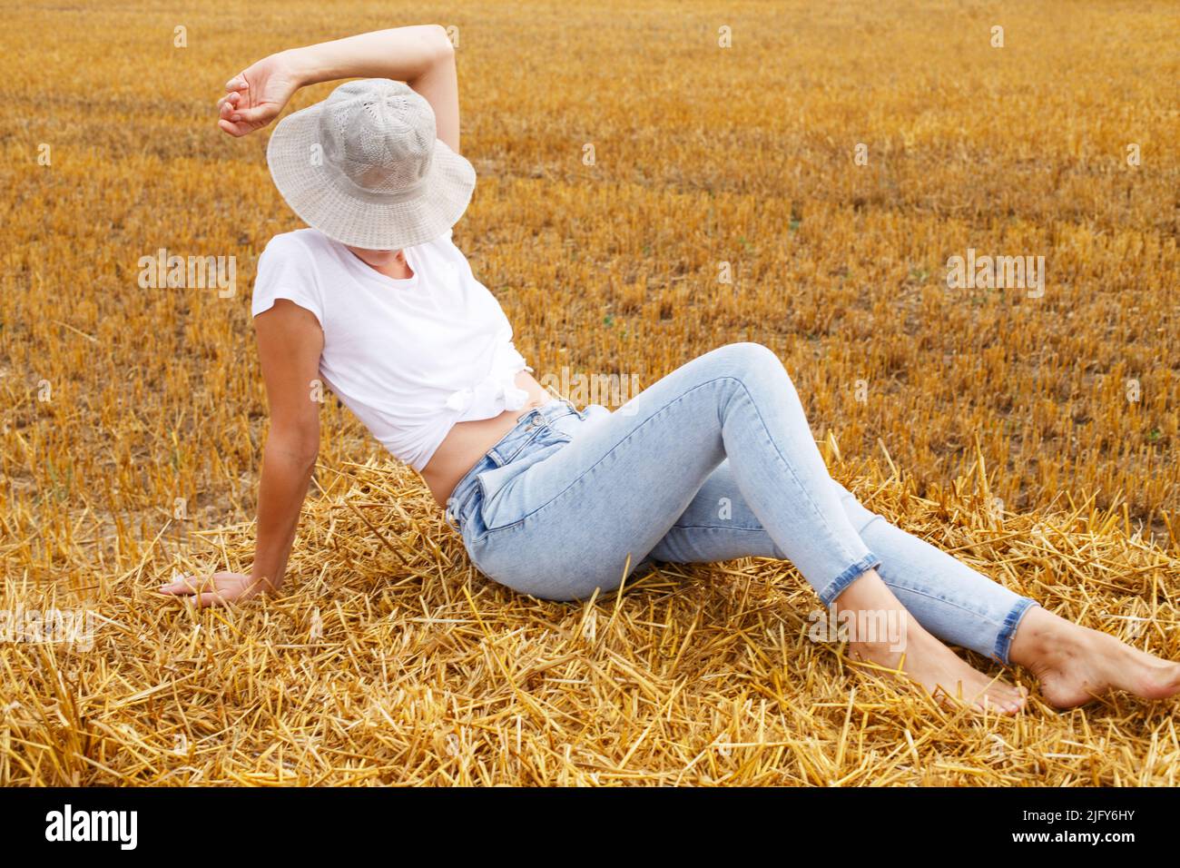 fille pieds nus avec chapeau de paille assis sur une botte de foin sur une balle dans le champ agricole après la récolte. Banque D'Images