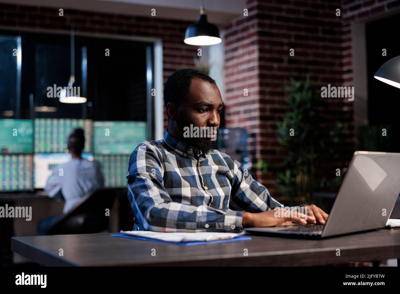 Homme d'affaires afro-américain assis à un bureau dans un espace de travail tout en utilisant un ordinateur portable pour analyser la tendance du marché. Trader professionnel faisant l'analyse technique concernant le commerce de stock de forex la nuit. Banque D'Images