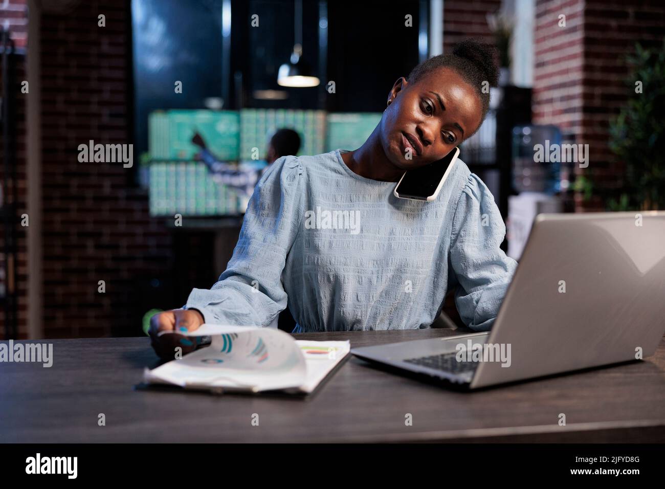 Agent de négociation de la bourse afro-américaine de forex au téléphone avec un investisseur parlant de risques financiers. Analyste de la société d'investissement assis dans l'espace de travail la nuit pendant un appel mobile avec le client. Banque D'Images