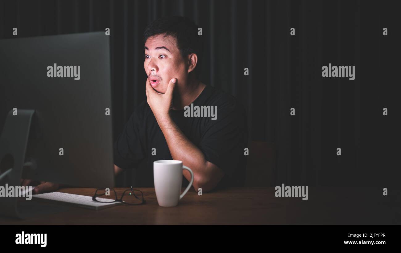 Début de travail dans le concept du matin : homme asiatique travaillant sur ordinateur dans la pièce sombre et regardant à l'écran avec le visage passionnant. Espace sombre pour le texte an Banque D'Images