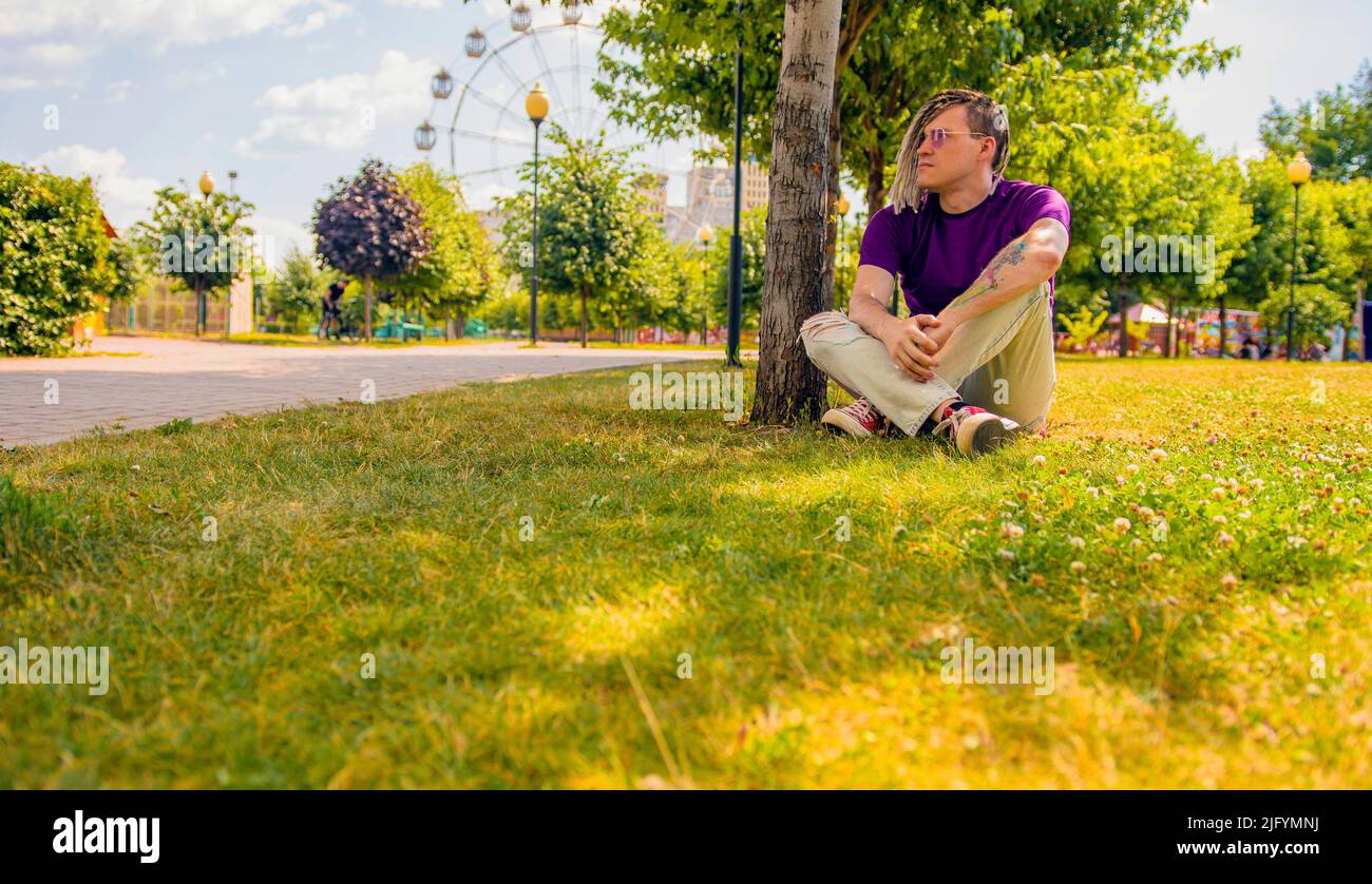 Un bel homme blanc avec des dreadlocks dans des vêtements décontractés, assis dans le parc sur l'herbe et regardant soigneusement dans le loin. Un hipster reposant Banque D'Images