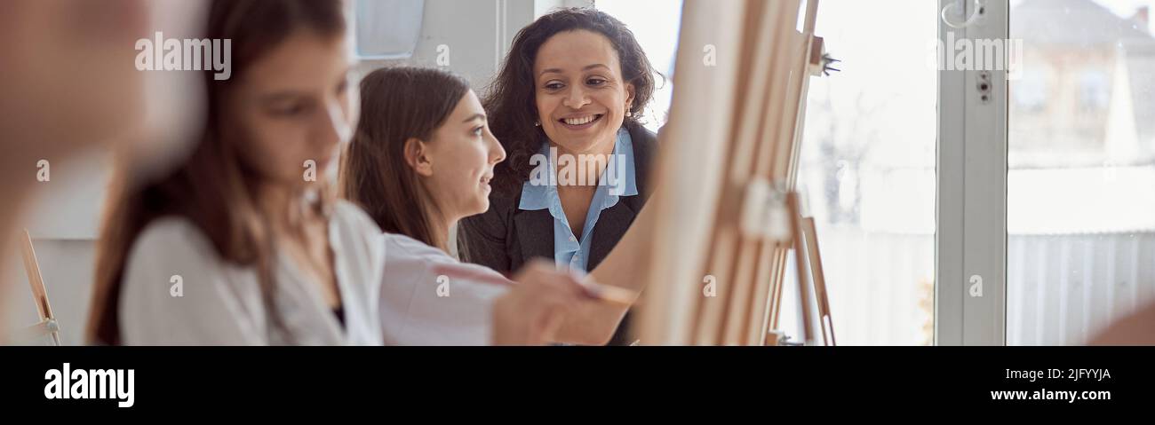 Jeune beau professeur confiant aide un enfant à tirer parti d'une leçon de groupe dans une salle de classe minimaliste moderne blanche Banque D'Images