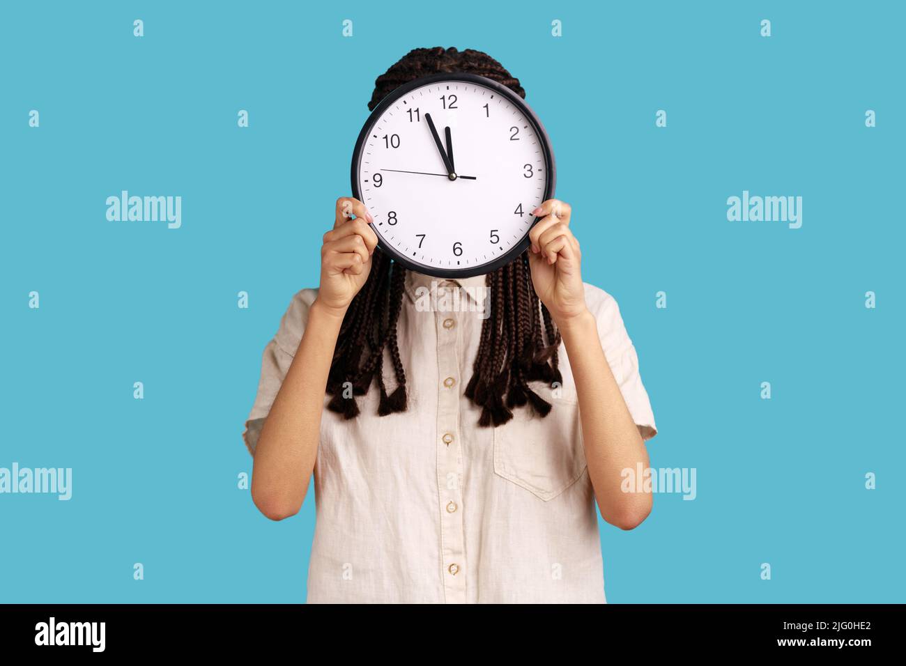 Temps de travail. Femme avec des dreadlocks se cachant face derrière une grande horloge murale, gestion du temps, rendez-vous de réunion d'affaires et de calendrier, portant une chemise blanche. Studio d'intérieur isolé sur fond bleu. Banque D'Images