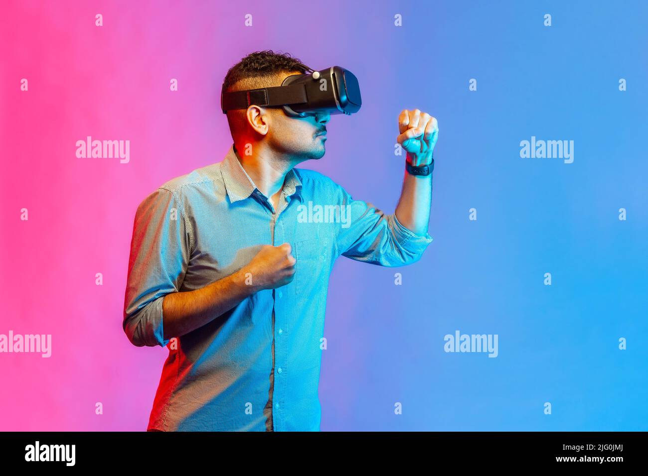 Homme en chemise dans des lunettes de réalité virtuelle à la tête jouant au jeu de combat, tenant les poings serrés prêts à la boxe, à l'aide d'un casque VR. Studio d'intérieur isolé sur fond de néon coloré. Banque D'Images