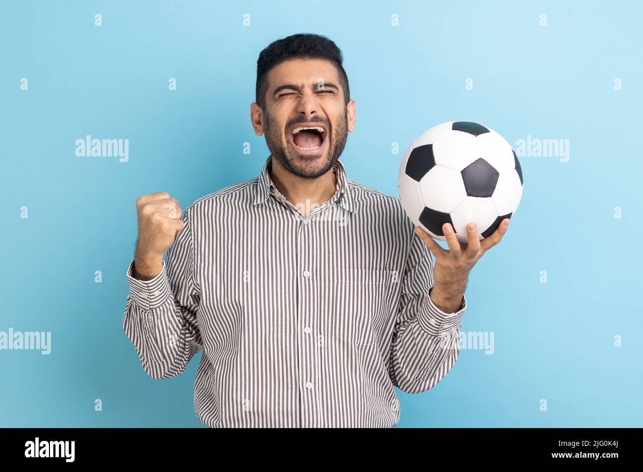 Un homme d'affaires barbu criait largement en bouche, célébrant la victoire de l'équipe de football préférée, tenant le ballon dans les mains et portant une chemise rayée. Studio d'intérieur isolé sur fond bleu. Banque D'Images