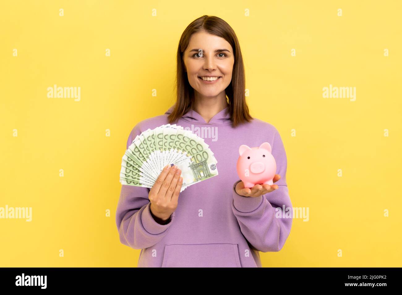 Souriante femme positive avec cheveux foncés tenant une grande somme d'argent et de la banque de porc, investissement rentable, portant le chandails pourpre. Studio d'intérieur isolé sur fond jaune. Banque D'Images