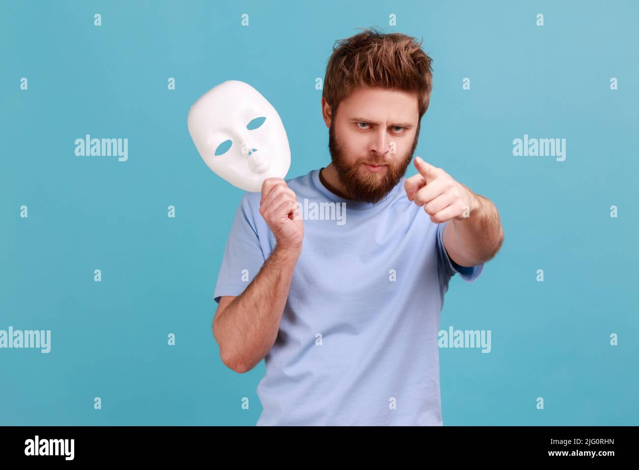 Portrait d'un jeune homme barbu adulte agressif et strict tenant le masque blanc à la main, pointant le doigt vers l'appareil photo avec une expression bossy sérieuse. Studio d'intérieur isolé sur fond bleu. Banque D'Images