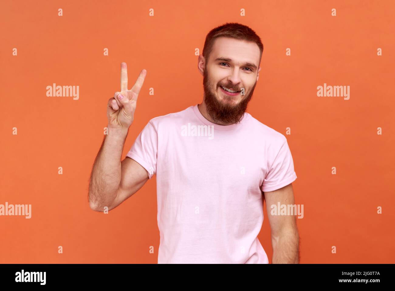 Portrait d'un jeune homme sympathique et barbu adulte debout avec une main levée et montrant le signe v ou la paix, le geste de la victoire, portant un T-shirt rose. Studio d'intérieur isolé sur fond orange. Banque D'Images