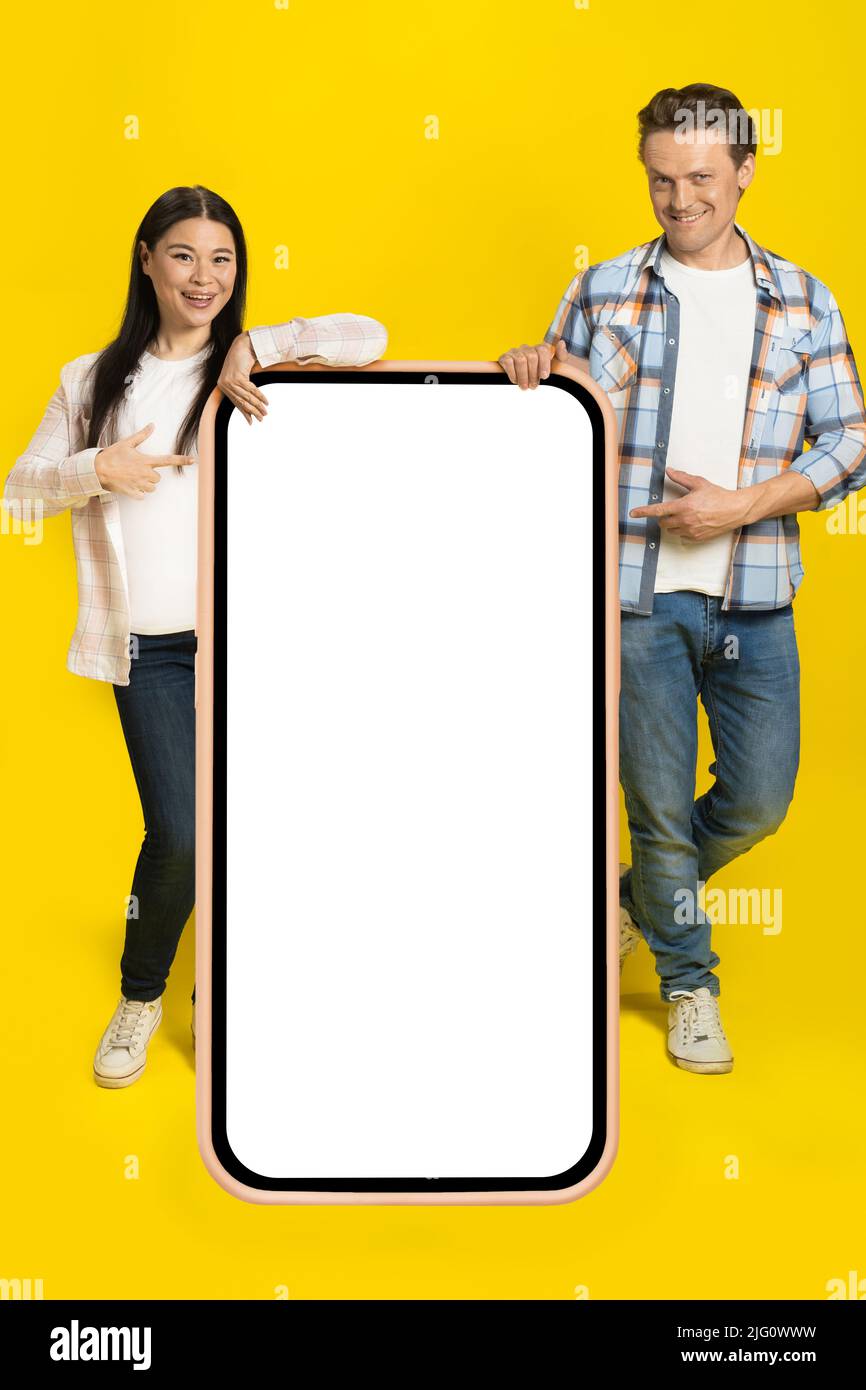 Pointant vers l'écran blanc homme caucasien et femme asiatique debout adossé sur un immense smartphone avec écran blanc, publicité d'application mobile isolée sur fond jaune. Positionnement du produit. Banque D'Images