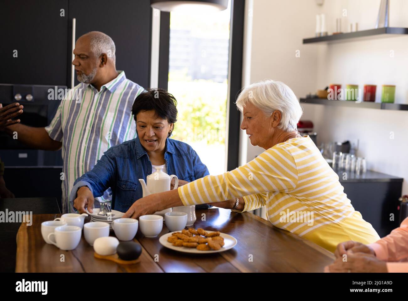 Des femmes âgées biraciales servent du café et des biscuits à des amis sur une table en bois dans une maison de soins infirmiers Banque D'Images