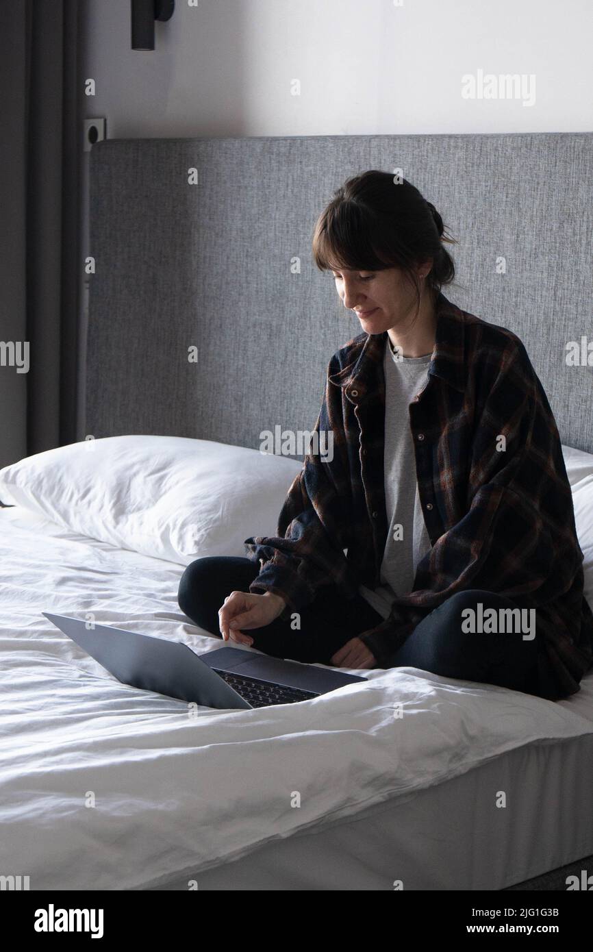Une jeune femme s'assoit sur le lit avec un ordinateur portable à la maison et communique agréablement avec ses amis et sa famille ou passe du temps à regarder un film. Banque D'Images