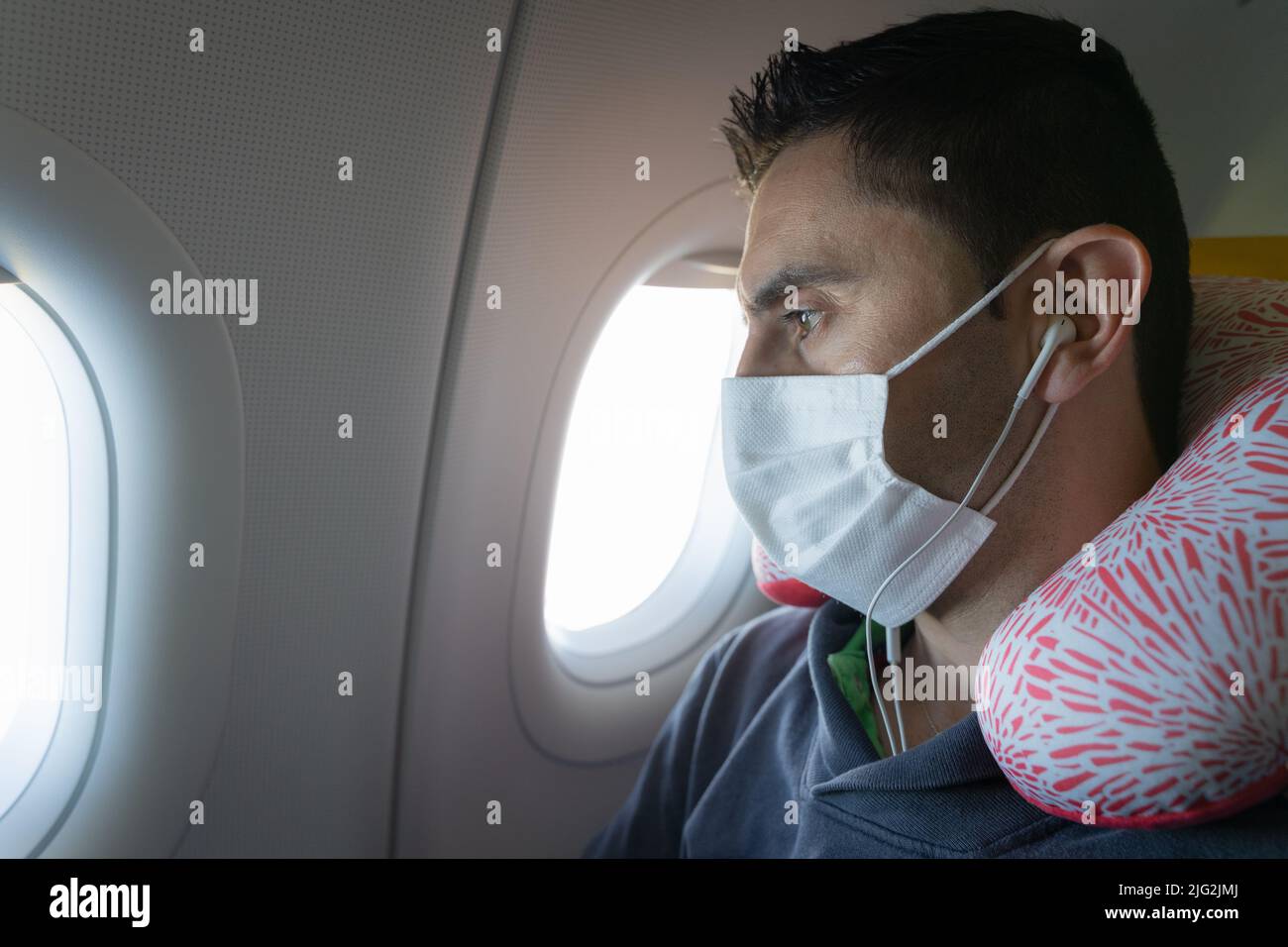 Homme avec masque, écouteurs et oreiller de cou regardant à travers la fenêtre plane. Mesures de protection de la santé des avions, concepts de confort long voyage Banque D'Images