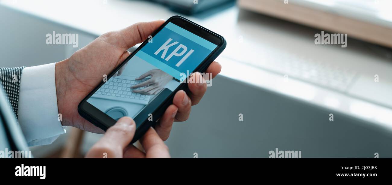 Homme au bureau, portant une chemise et une veste, tenant son smartphone, qui lit le texte KPI, pour indicateur de performance clé, dans son écran, dans un panor Banque D'Images