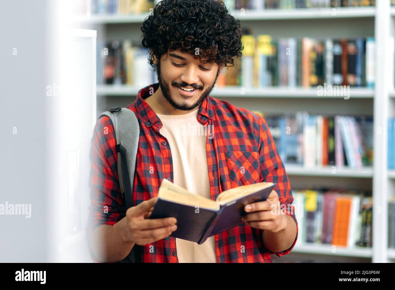 Spécialisé, intelligent et positif étudiant indien ou arabe d'université ou d'université, debout dans une bibliothèque près de la bibliothèque, la lecture d'un livre intéressant, la préparation à l'examen, faire des devoirs, sourire Banque D'Images