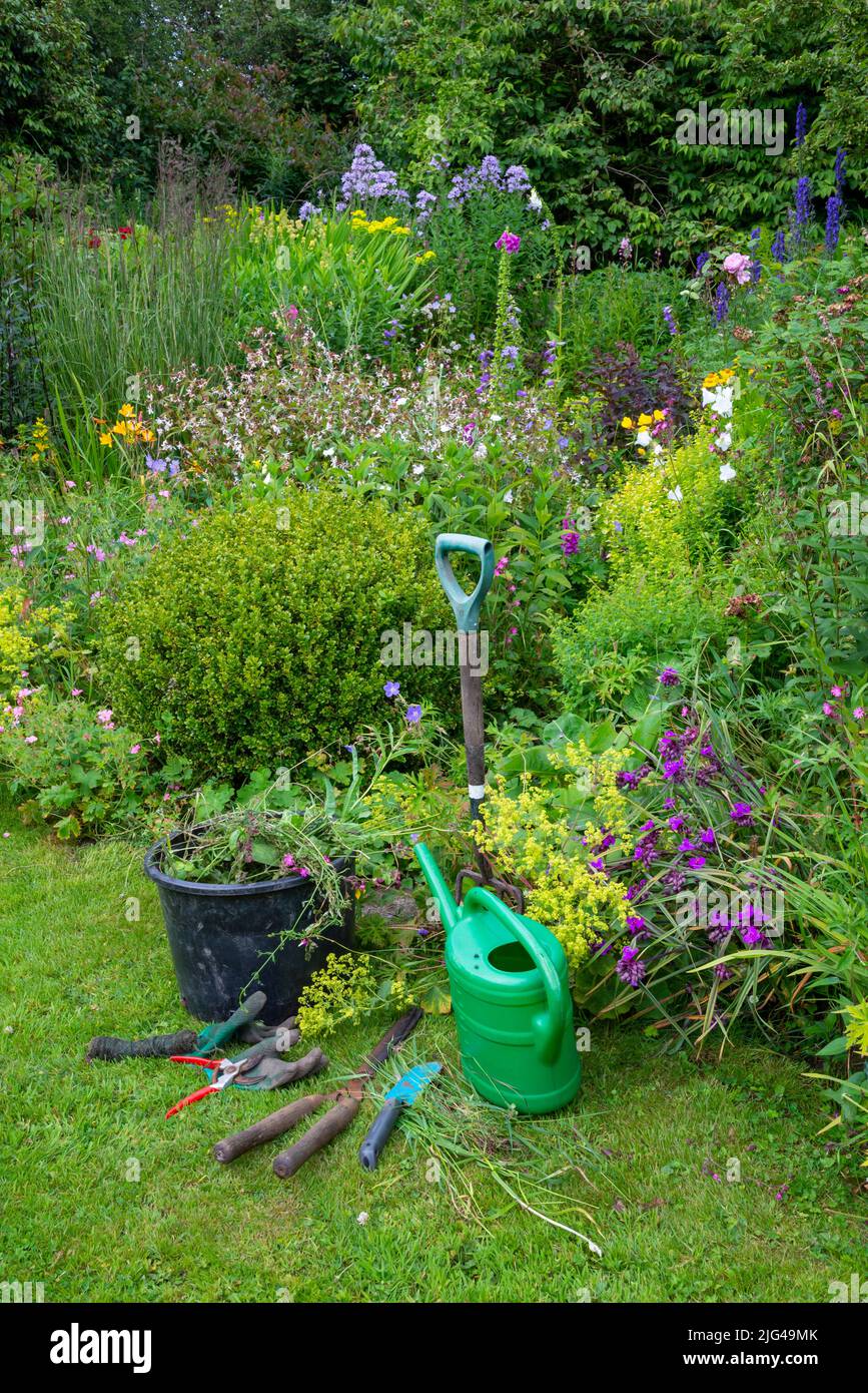 Jardin anglais de campagne début juillet. Une sélection d'outils bien utilisés à côté des frontières pleines de diverses plantes. Banque D'Images