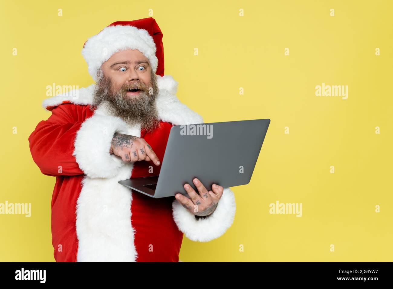 le père noël en surpoids stupéfié pointe vers un ordinateur portable isolé sur du jaune Banque D'Images
