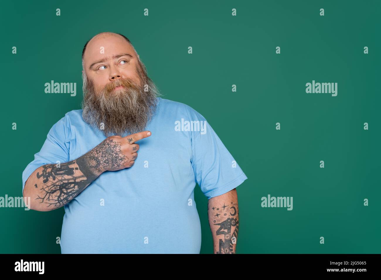 homme de surpoids réfléchi avec tatouages pointant avec le doigt et regardant loin isolé sur le vert Banque D'Images
