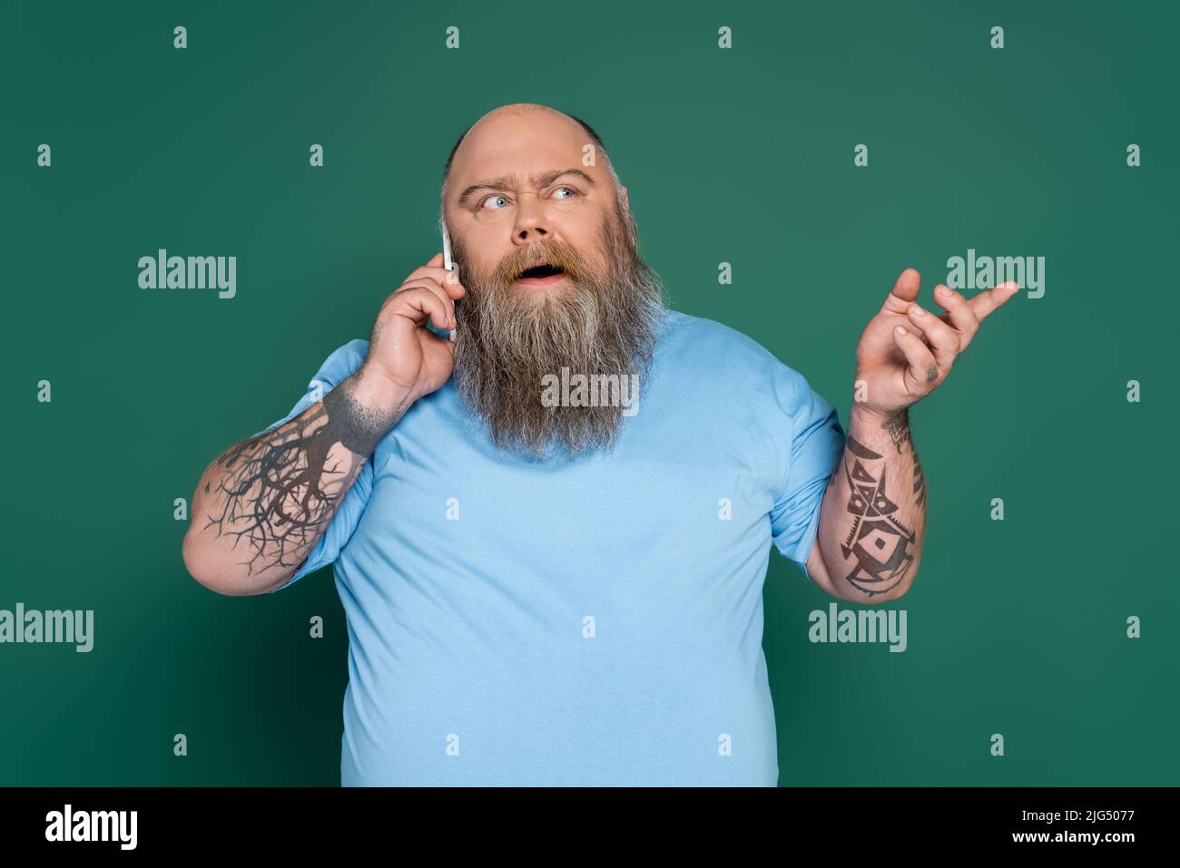 homme sérieux en surpoids avec tatouages parlant sur smartphone et pointant avec le doigt isolé sur le vert Banque D'Images