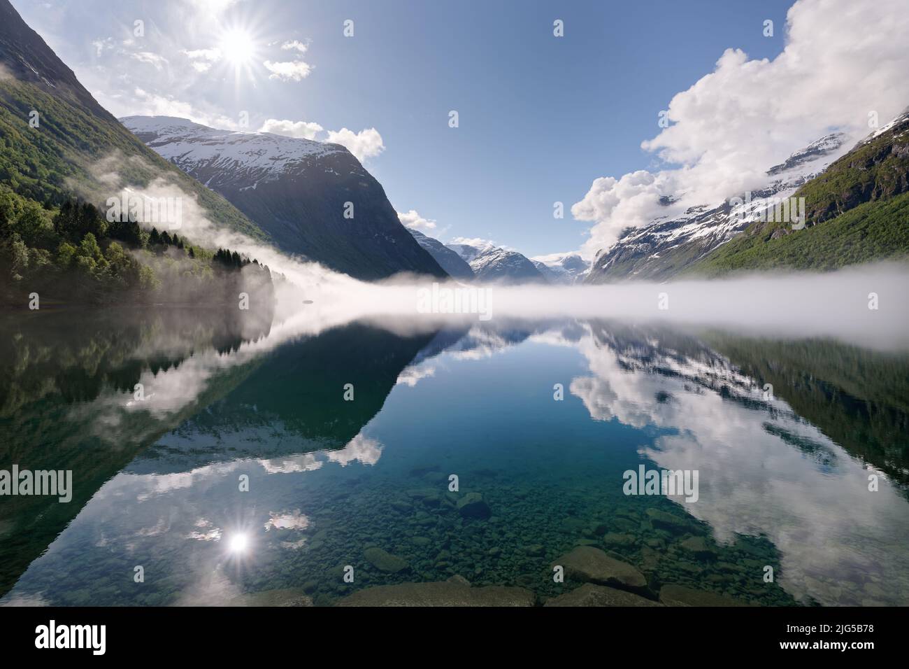 Vue sur un lac entouré de hautes montagnes partiellement enneigées en Norvège, le soleil vient de devenir visible, une banque de brouillard se trouve sur le lac, reflet Banque D'Images