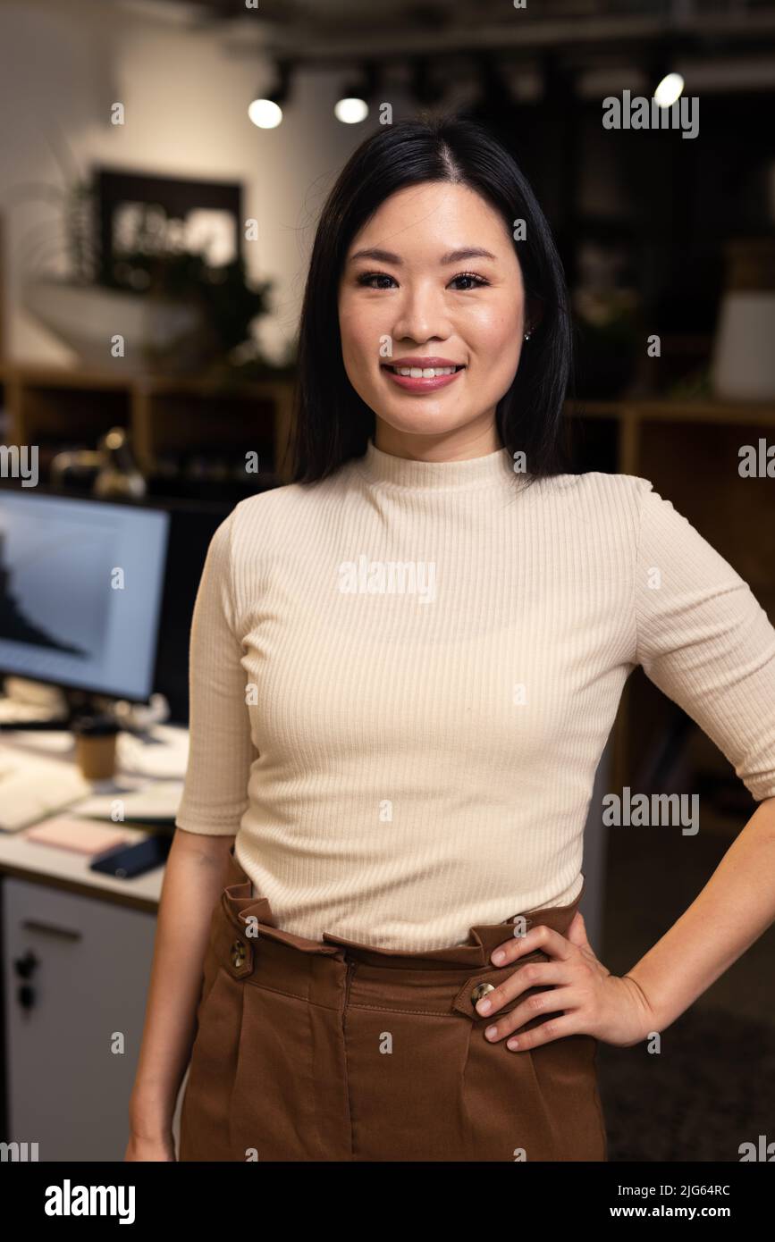 Portrait d'une femme asiatique souriante, main sur la hanche, debout dans un bureau moderne la nuit Banque D'Images