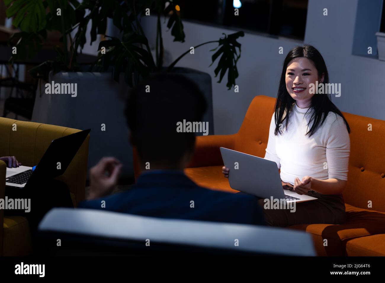 Femme d'affaires asiatique souriante communiquant avec un collègue tout en travaillant sur un ordinateur portable sur un canapé Banque D'Images
