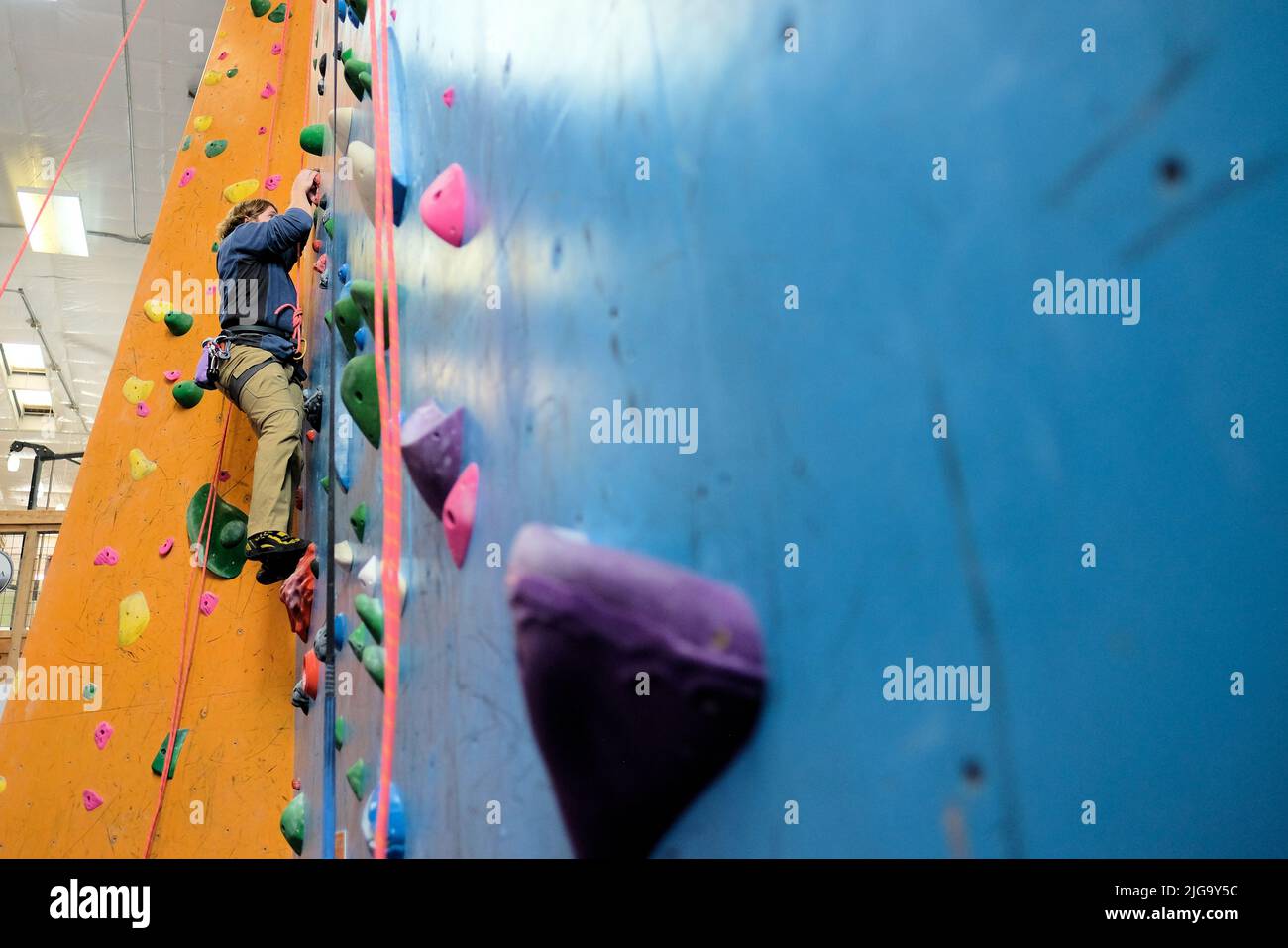 Jeune homme grimpant sur un mur d'escalade intérieur vu d'en-dessous ; cordes et cales d'escalade pour le soutien de l'ascension. Banque D'Images